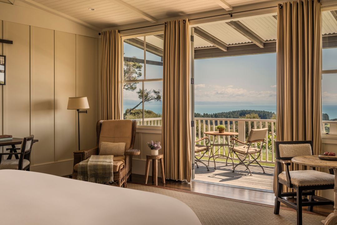 A hotel suite with neutral interiors, armchair, table with fresh fruit, desk, balcony and ocean views.